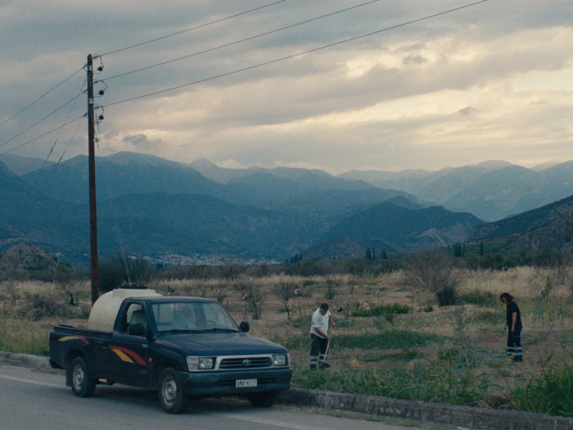 Pokrajinski prizor prikazuje temen pickup parkiran ob cesti, v bližini pa dva moška z orodjem urejata zelenico. V ozadju se razprostira veriga modrikastih gora pod oblačnim nebom. Slika daje občutek miru in rahle melanholije.