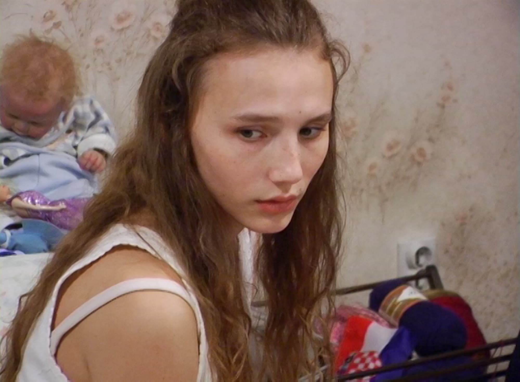 Dekle z dolgimi rjavimi lasmi sedi v notranjosti sobe in gleda zamišljeno v stran. V ozadju so igrače in dojenčkova lutka na postelji, stena pa ima nežne vzorce. Vzdušje je tiho in melanholično.