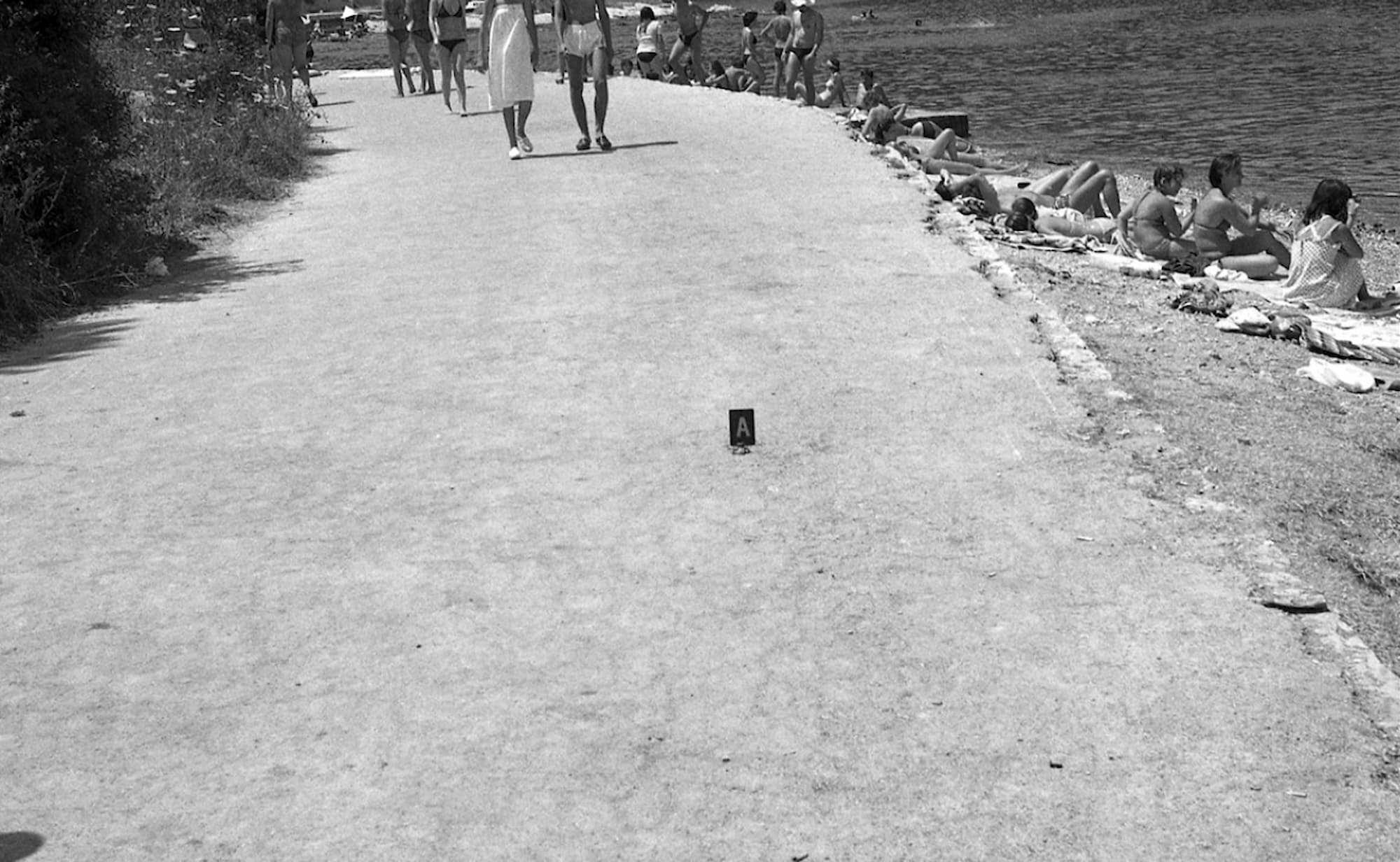 Črno-bela fotografija ob plaži: sprehajalci hodijo po potki, medtem ko se na desni strani ljudje sončijo. Na tleh stoji oznaka z velikim črnim A, ki nakazuje na raziskavo ali dogodek.