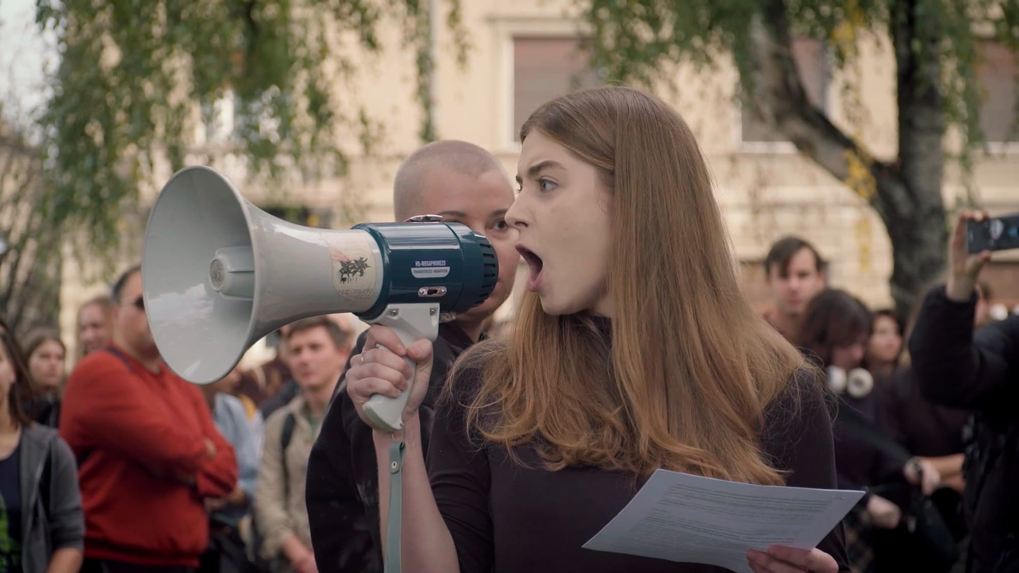 Mlada ženska govori skozi megafon pred množico ljudi na ulici. Prizor ujame energijo protesta ali javnega govora.