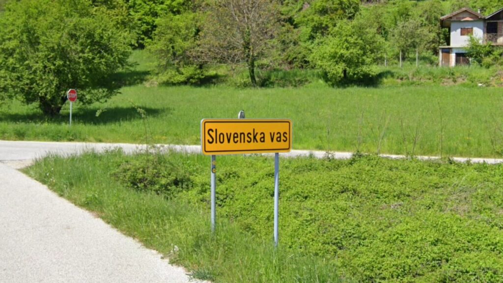 A yellow road sign with “Slovenska vas” stands by a rural road. Behind it is a green field with trees and a small house.