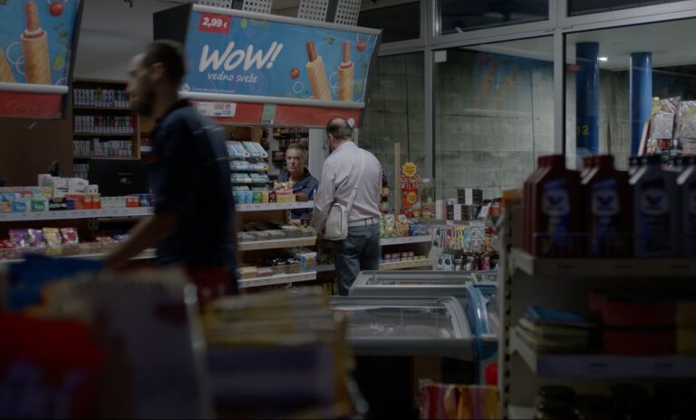 V trgovini moški v beli srajci govori s prodajalcem. V ospredju mimo gre druga oseba, ki je zamegljena.