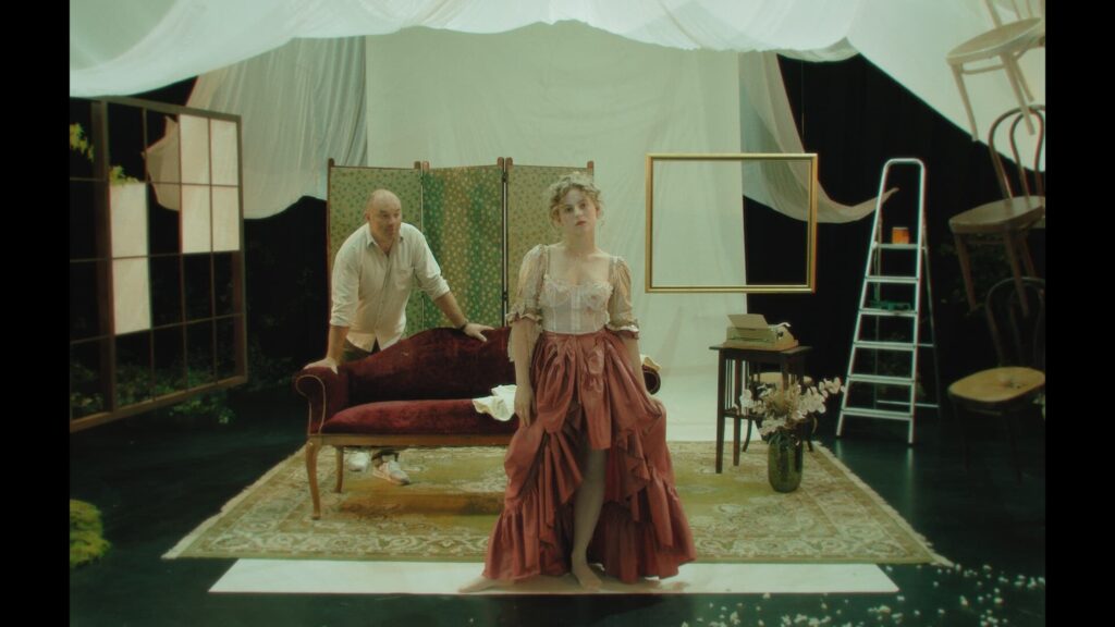 man and a woman are on a decorated stage, with vintage furniture and draped white cloths. The woman wears a historical dress, while the man stands behind a couch.