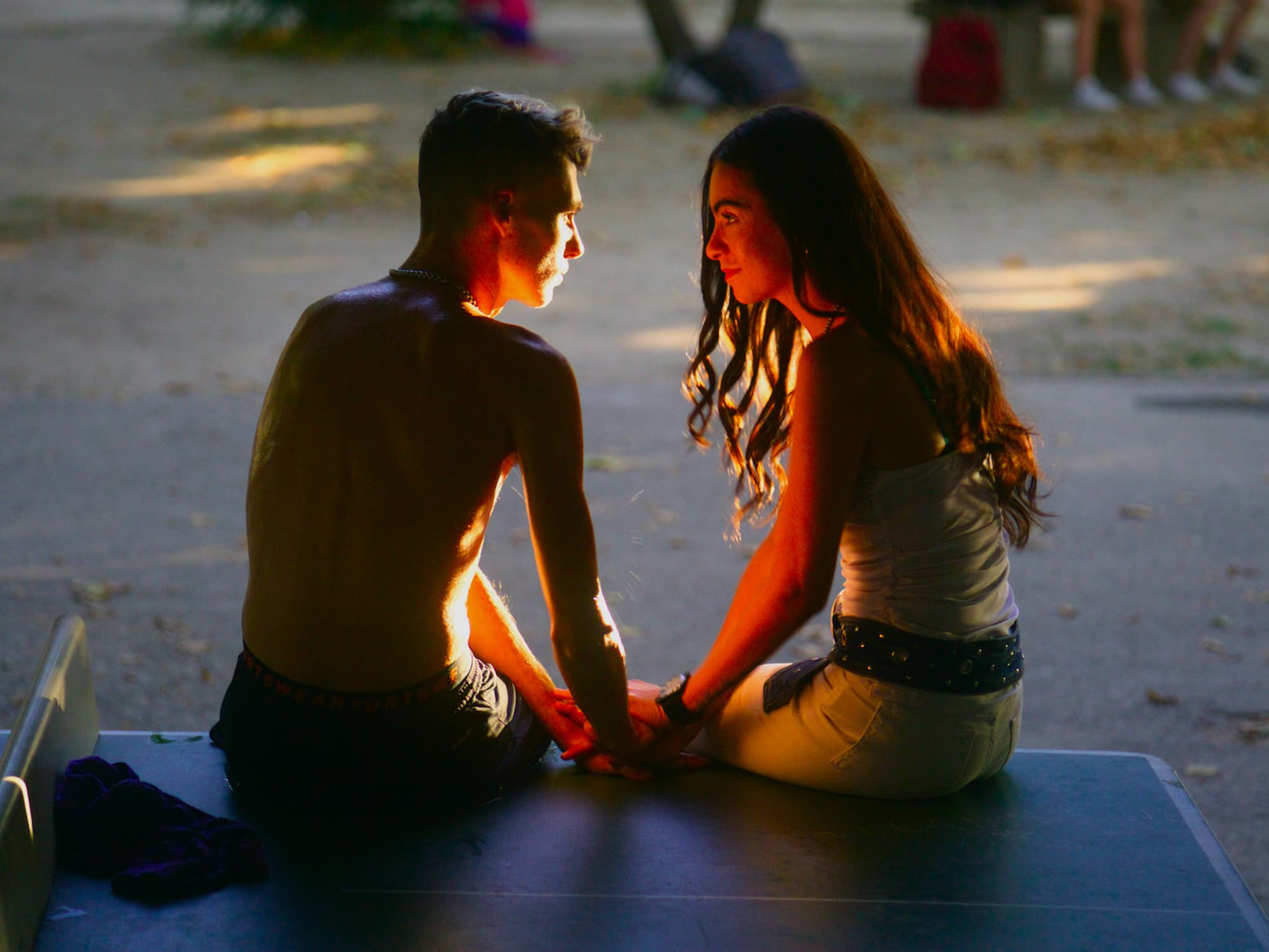 A shirtless boy and a girl sit facing each other on a table outdoors. They hold hands and look into each other's eyes, bathed in soft sunlight. The moment feels intimate and tender.