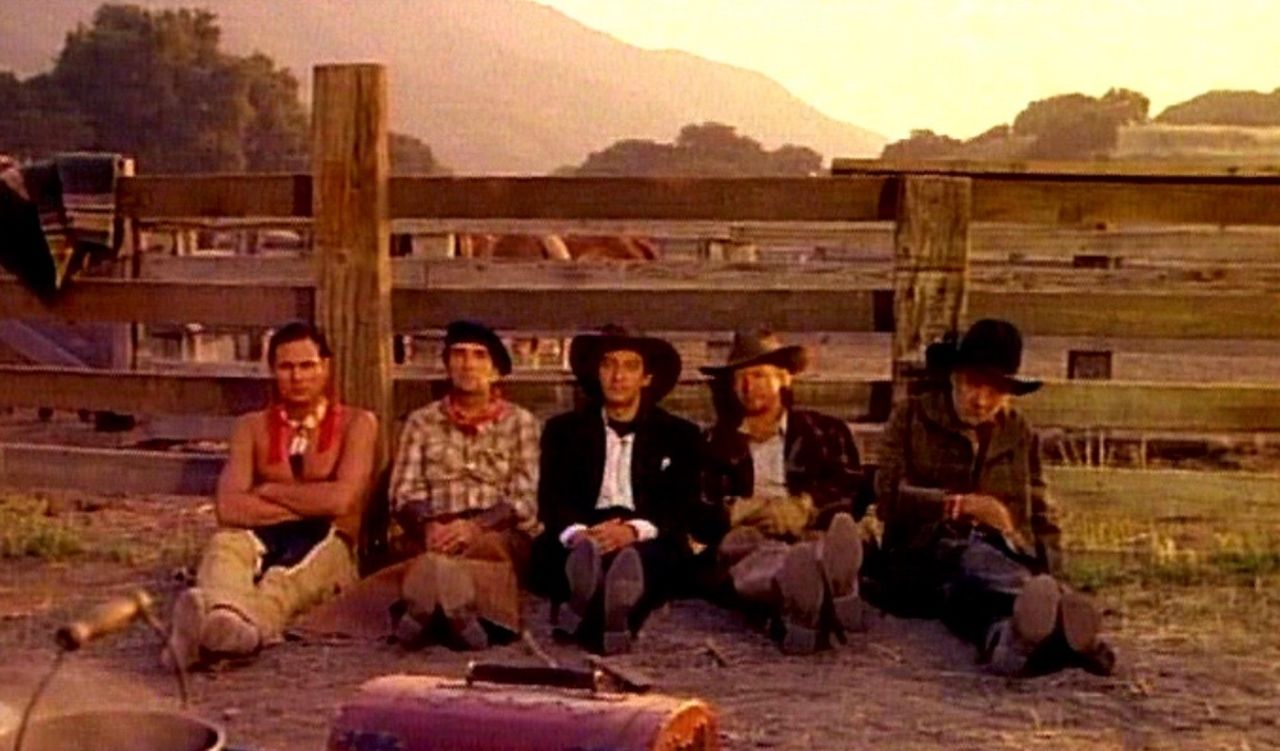 Prizor iz filma. Pet kavbojev sedi na tleh, naslonjeni so na leseno ograjo.