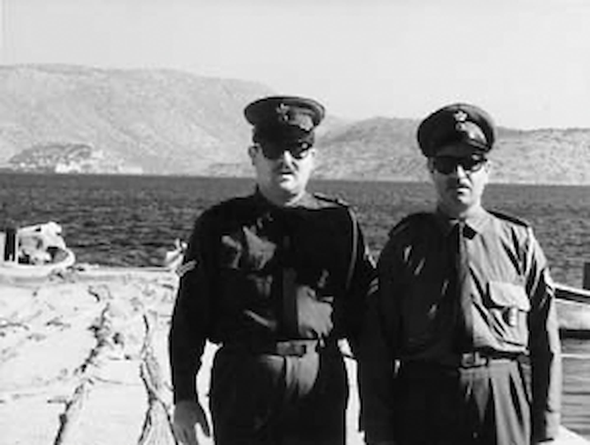 Prizor iz filma. Dva policaja ali oficirja stojita ob morju in gledata proti kameri.