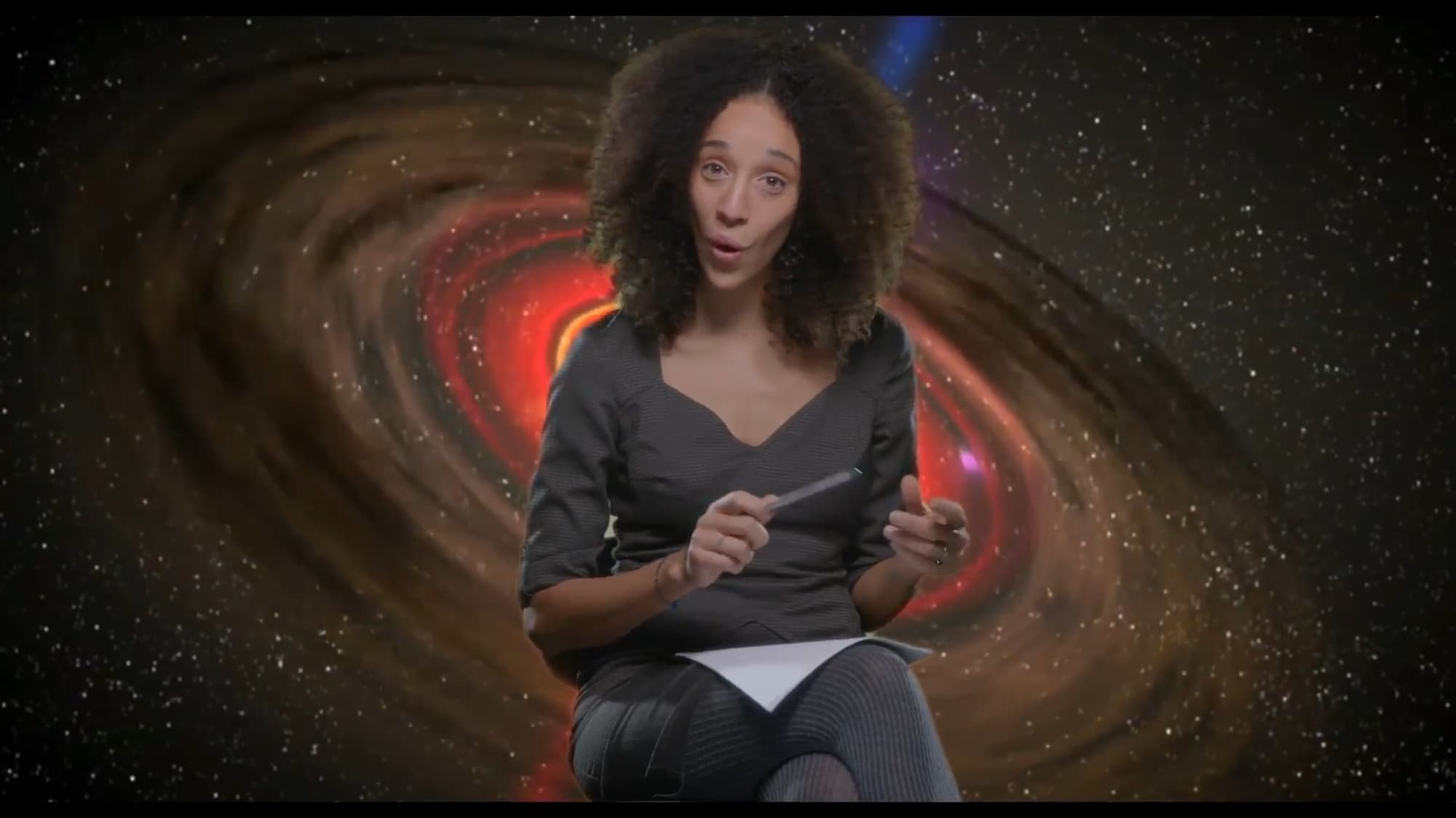 Prizor iz filma. Ženska sedi obrnjena proti kameri/gledalcu. Za njo je projekcija vesolja.